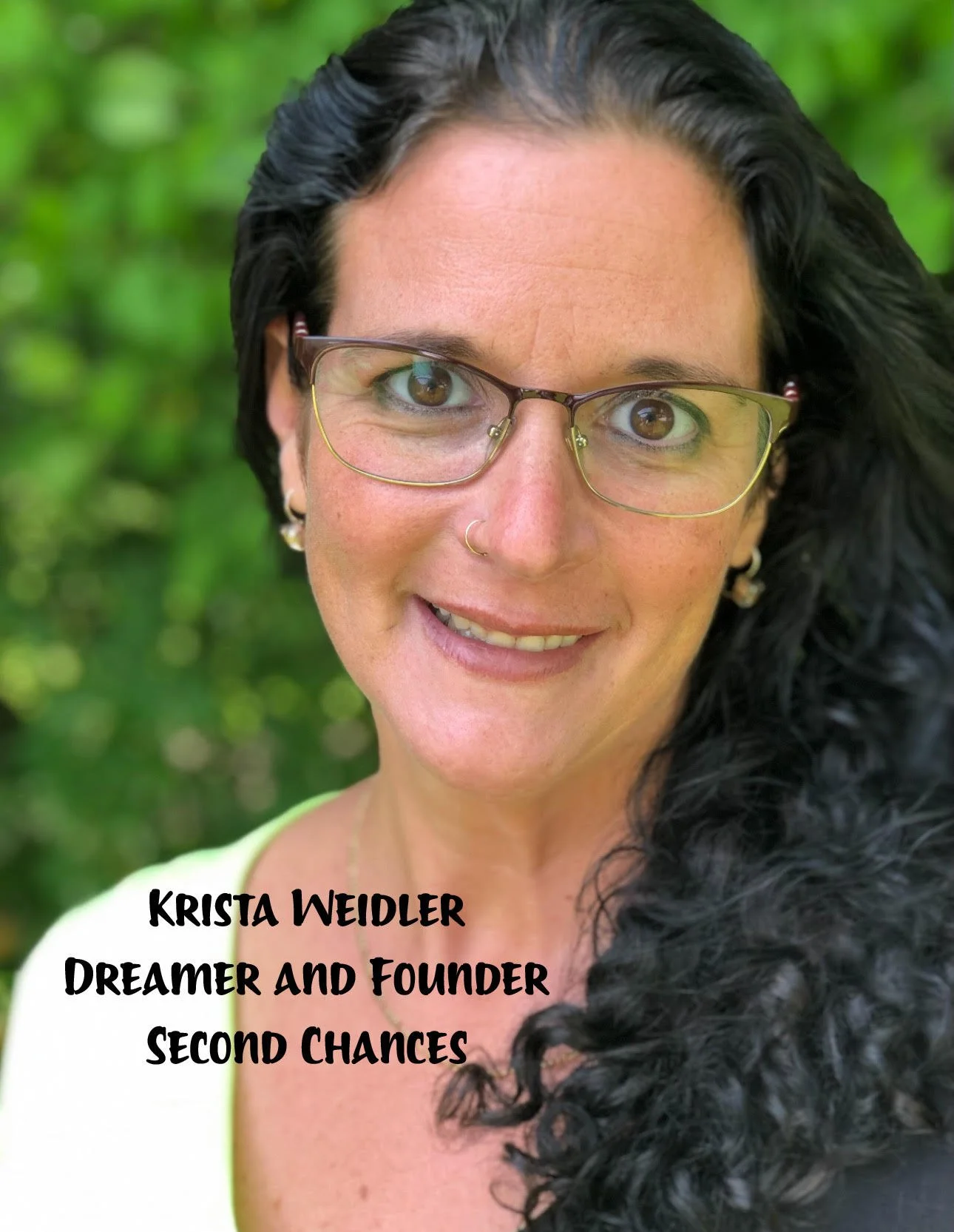 Close-up of a woman with long, curly black hair, glasses, and wearing a light-colored top, smiling in front of a green, leafy background. Text on the image reads: "Krista Weidler, Dreamer and Founder, Second Chances."