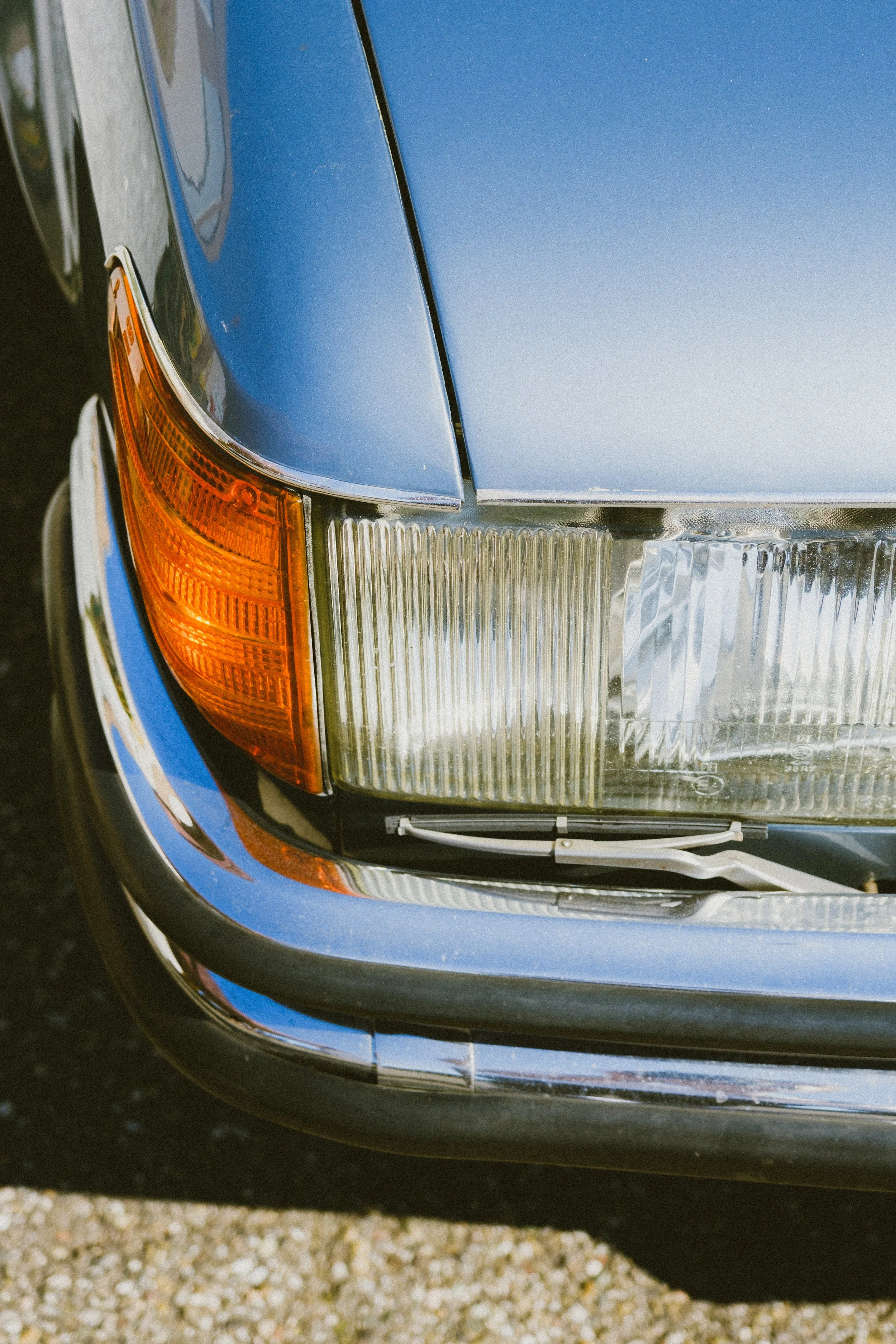 Close-up of the front left corner of a blue vintage car, showing part of the headlight, grille, and turn signal, parked on gravel.