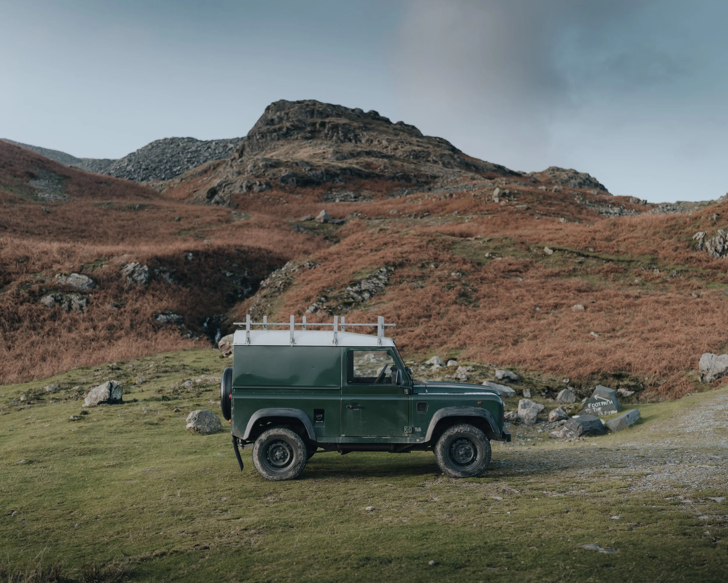 A green off-road vehicle parked on grassy terrain with hills and rocky formations in the background under a cloudy sky.