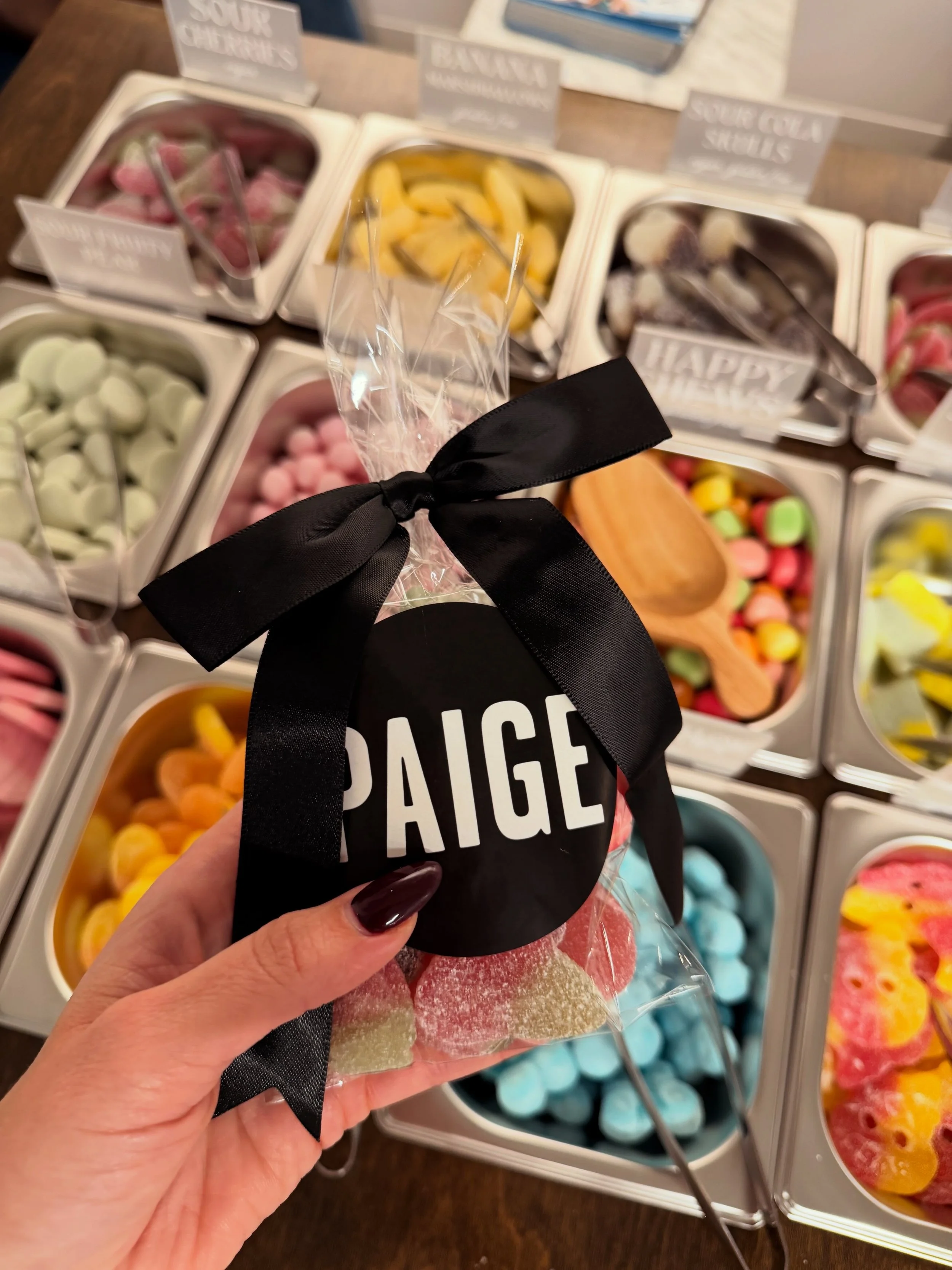 hand holding a bag of. candy with a black sticker that says paige in white lettering with black bow over a cart full of colorful candy