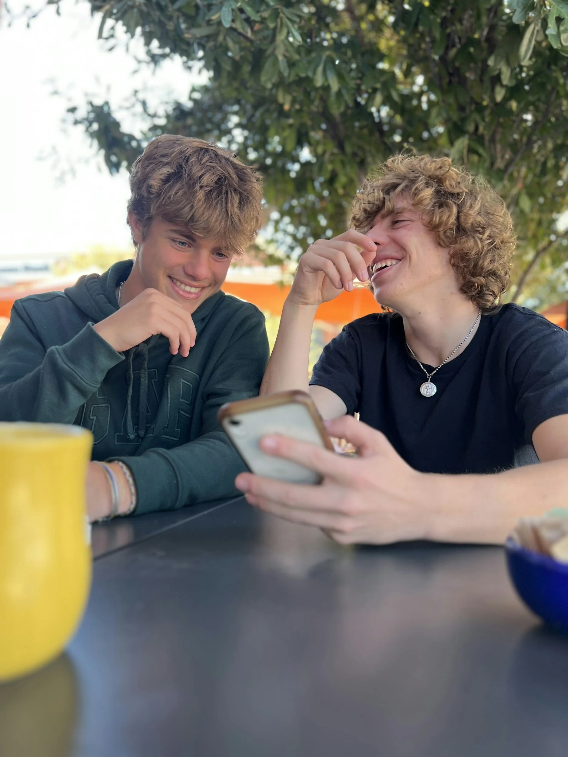 Two young men with curly hair sitting at a table outdoors, looking at a phone together and smiling, with colorful mugs in the foreground and trees in the background.