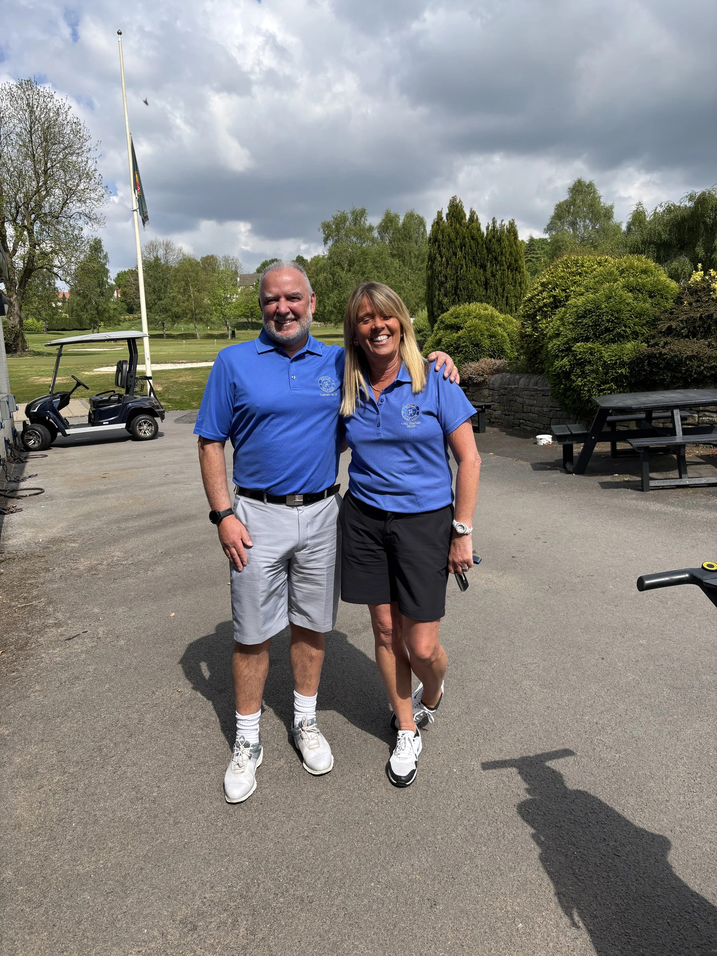 A man and woman standing outdoors at a golf course, smiling, wearing blue polo shirts, with trees, a golf cart, and a cloudy sky in the background.