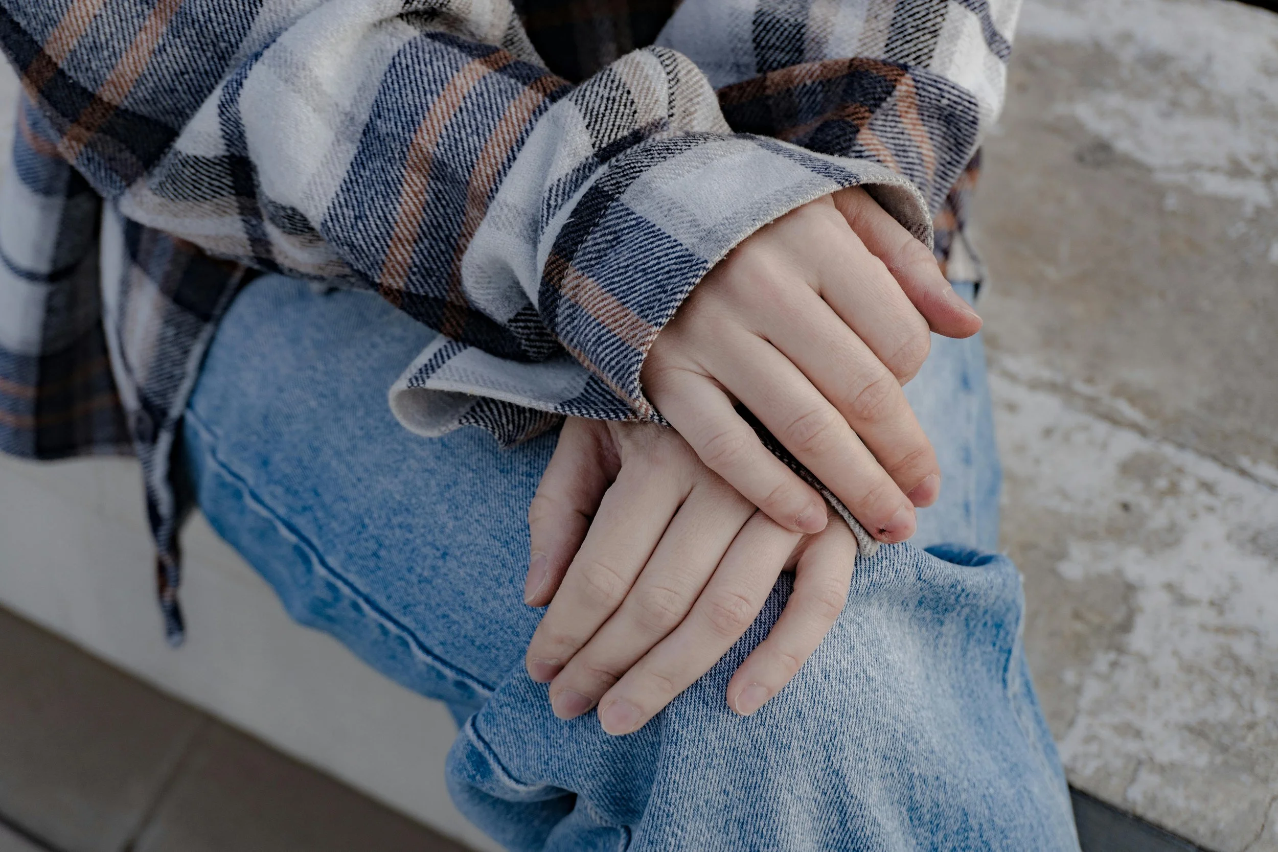 A close-up of two hands, one with a fair complexion and the other with a darker complexion. The person is sitting outdoors on a sandy surface, wearing casual clothing including a plaid shirt and jeans.