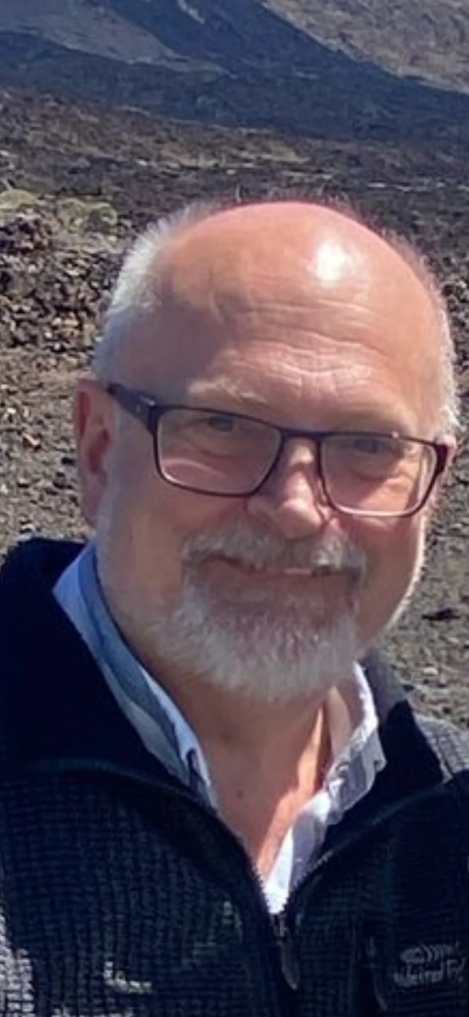 A smiling man with glasses and a beard standing outdoors in a rocky, mountainous landscape.