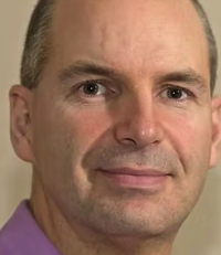 Close-up of a middle-aged man with short light-colored hair, wearing a light purple shirt, smiling softly.