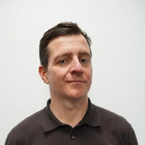 A man with short brown hair wearing a dark polo shirt standing against a white wall.