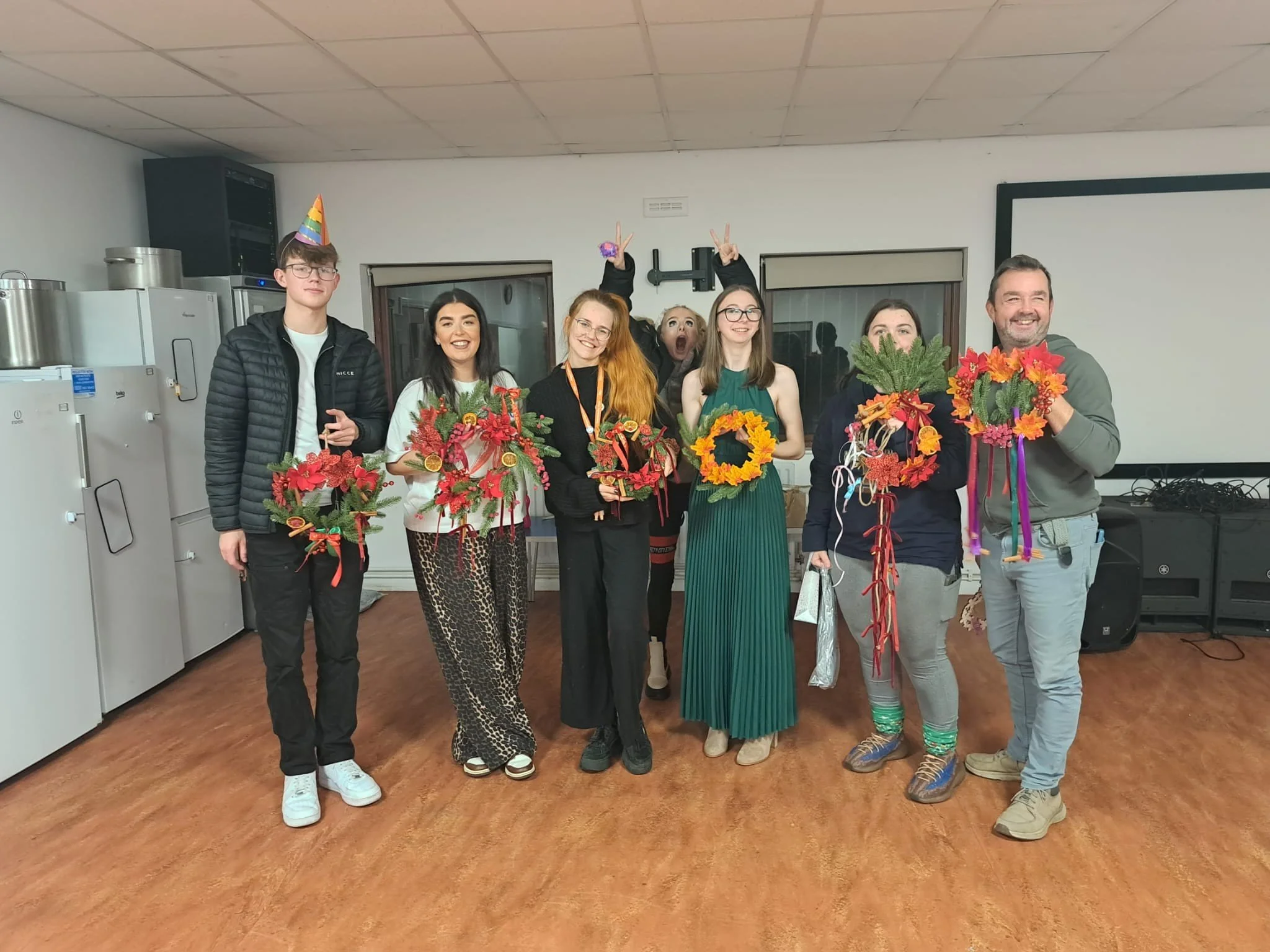 Group of people celebrating with party hats and festive decorations, holding floral arrangements, in an indoor setting.