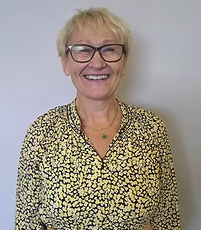 A woman with short blonde hair and glasses, smiling, wearing a patterned blouse, standing against a plain wall.
