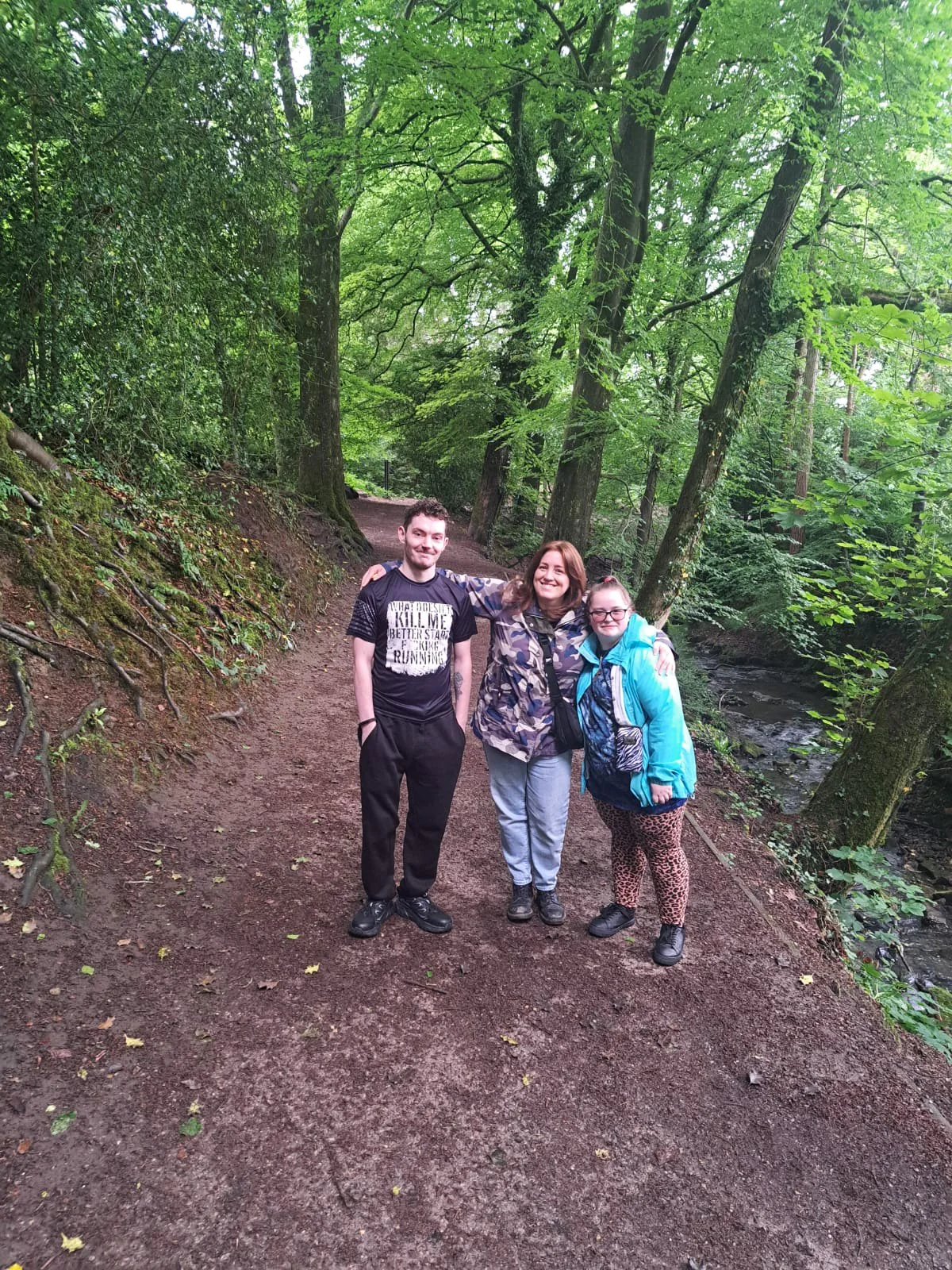 Three people standing on a forest trail, smiling, with trees and greenery around them.