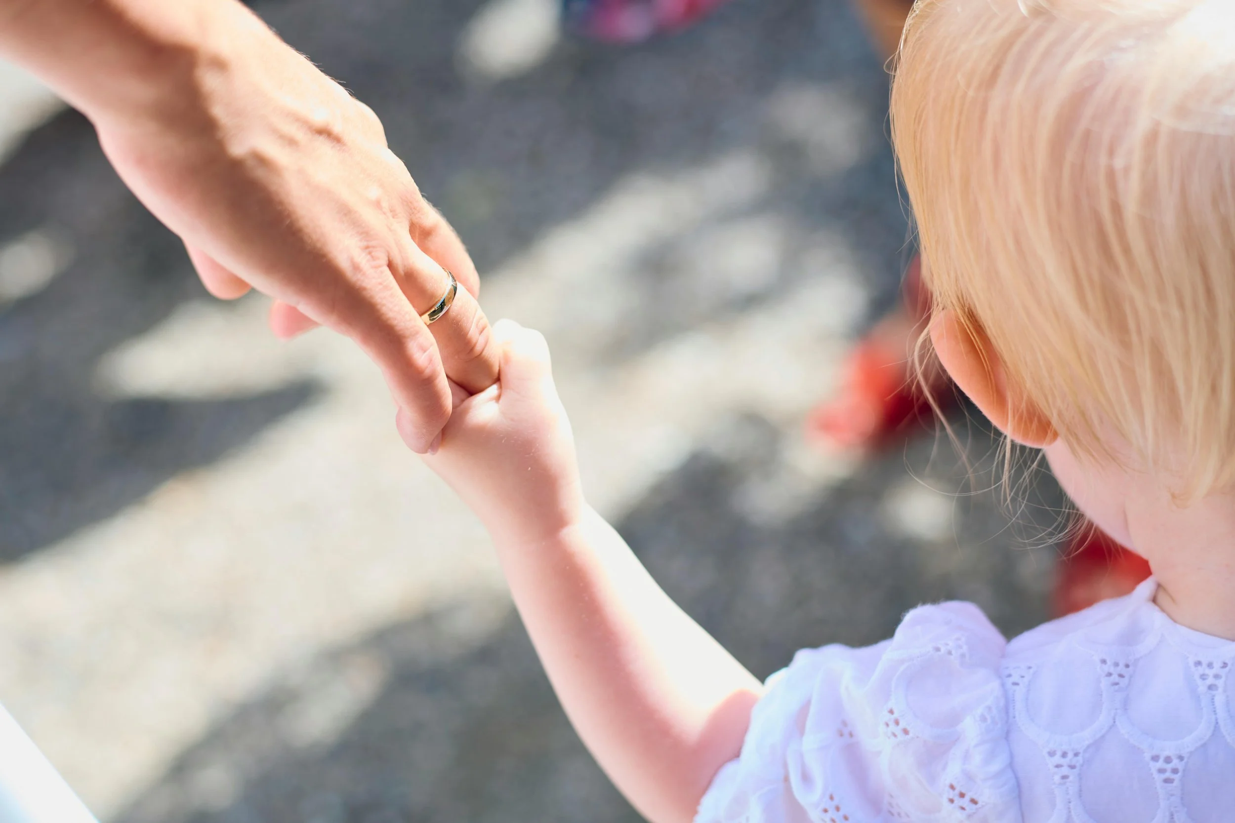 A young girl with blonde hair holding hands with an adult, outdoors on a sunny day.