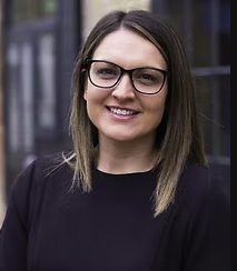 A woman with shoulder-length brown hair, glasses, and a black top, smiling outdoors with buildings in the background.
