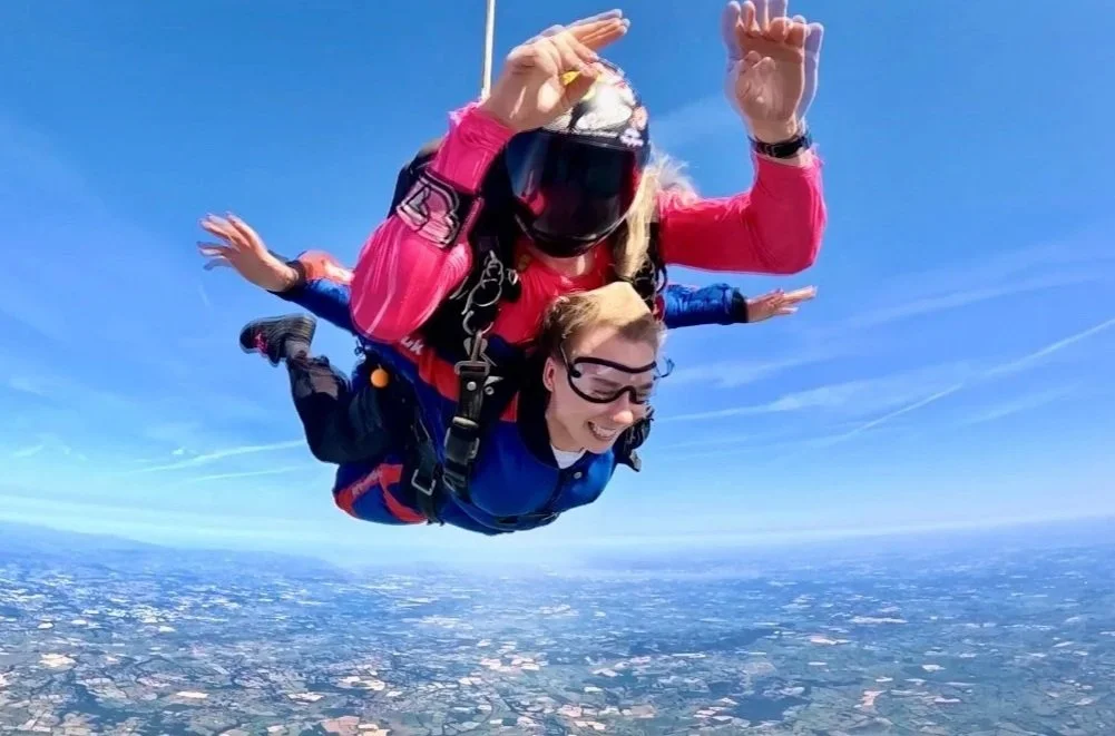 Two people tandem skydiving, free-falling with arms spread out over a landscape below on a clear day.