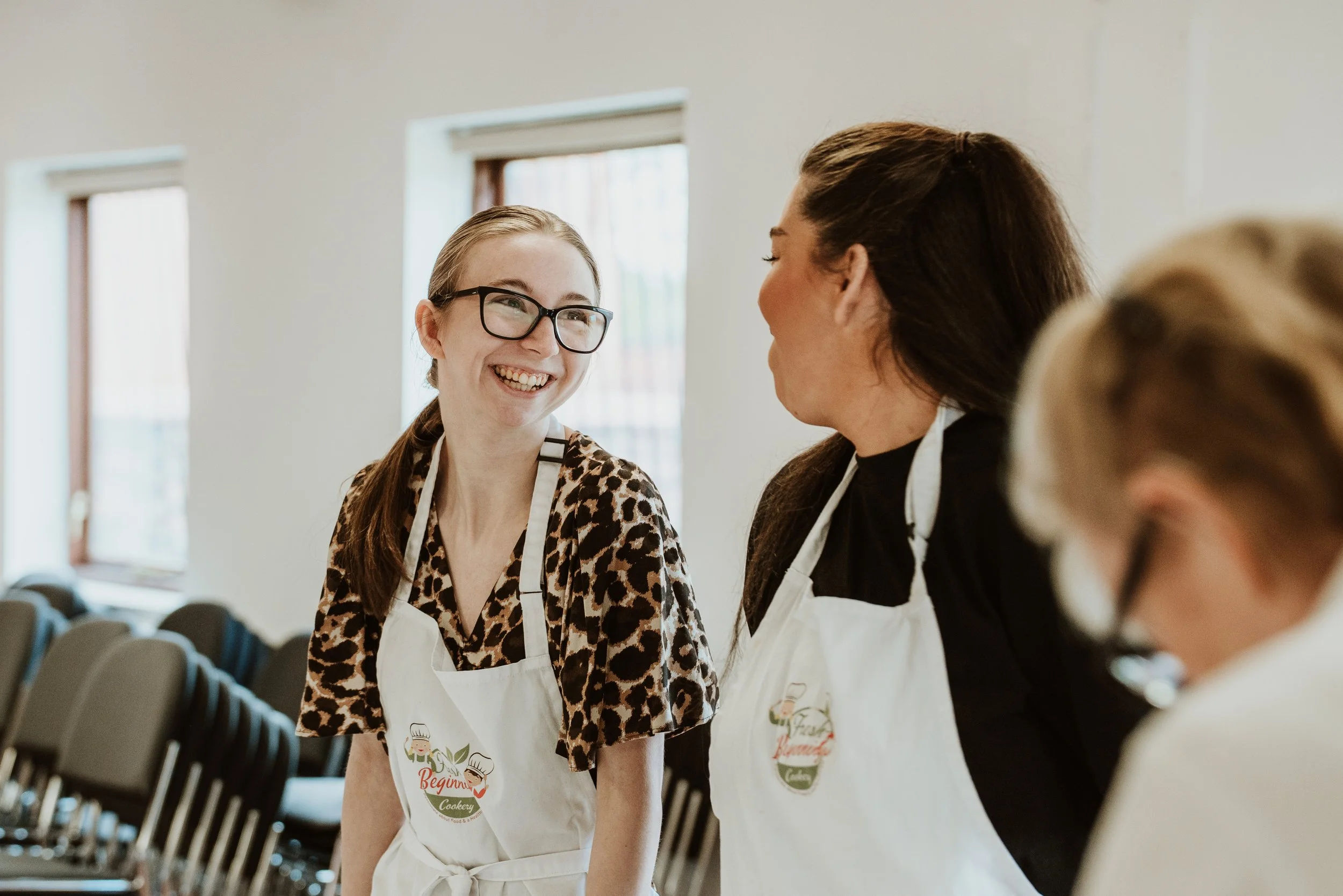 Two women, one with glasses and a leopard print top, and the other with dark hair, are smiling and talking in a room with large windows. Both are wearing white aprons with a logo.