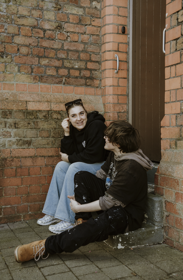 A young woman and a young man sitting on steps outside near a brick wall, smiling and chatting.