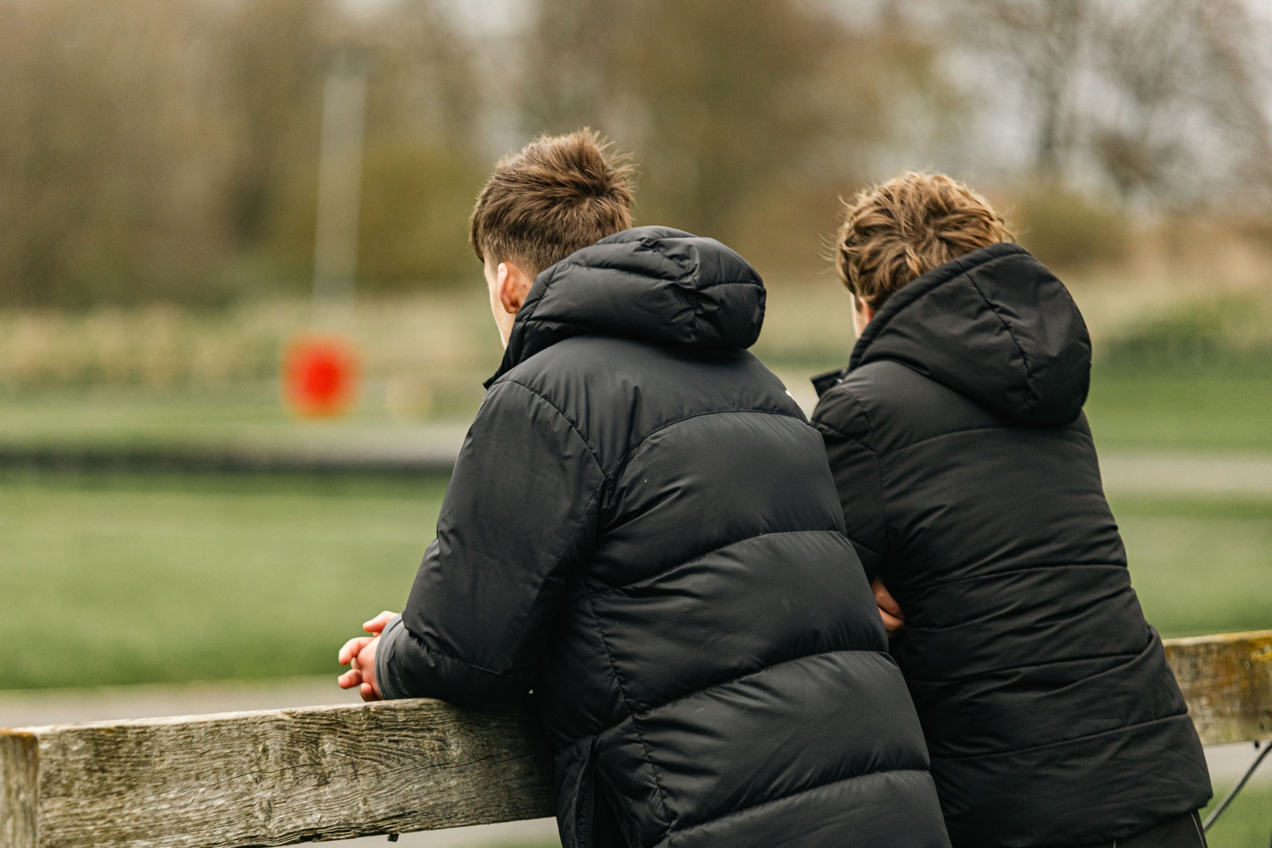 Two people, dressed warmly in black padded jackets with hoods, are sitting on a wooden log in an outdoor park or green space. They are sitting very close, with one person possibly resting their head on the other's shoulder, looking thoughtfully into the distance.