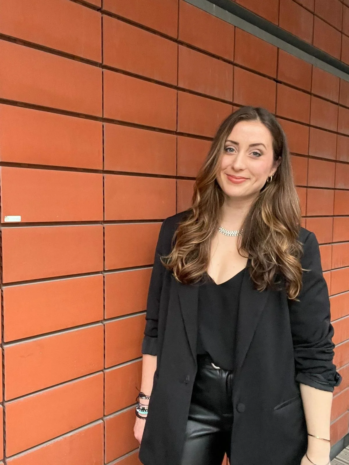 A woman with long wavy brown hair wearing a black blazer, black top, and black leather pants standing in front of a red brick wall.