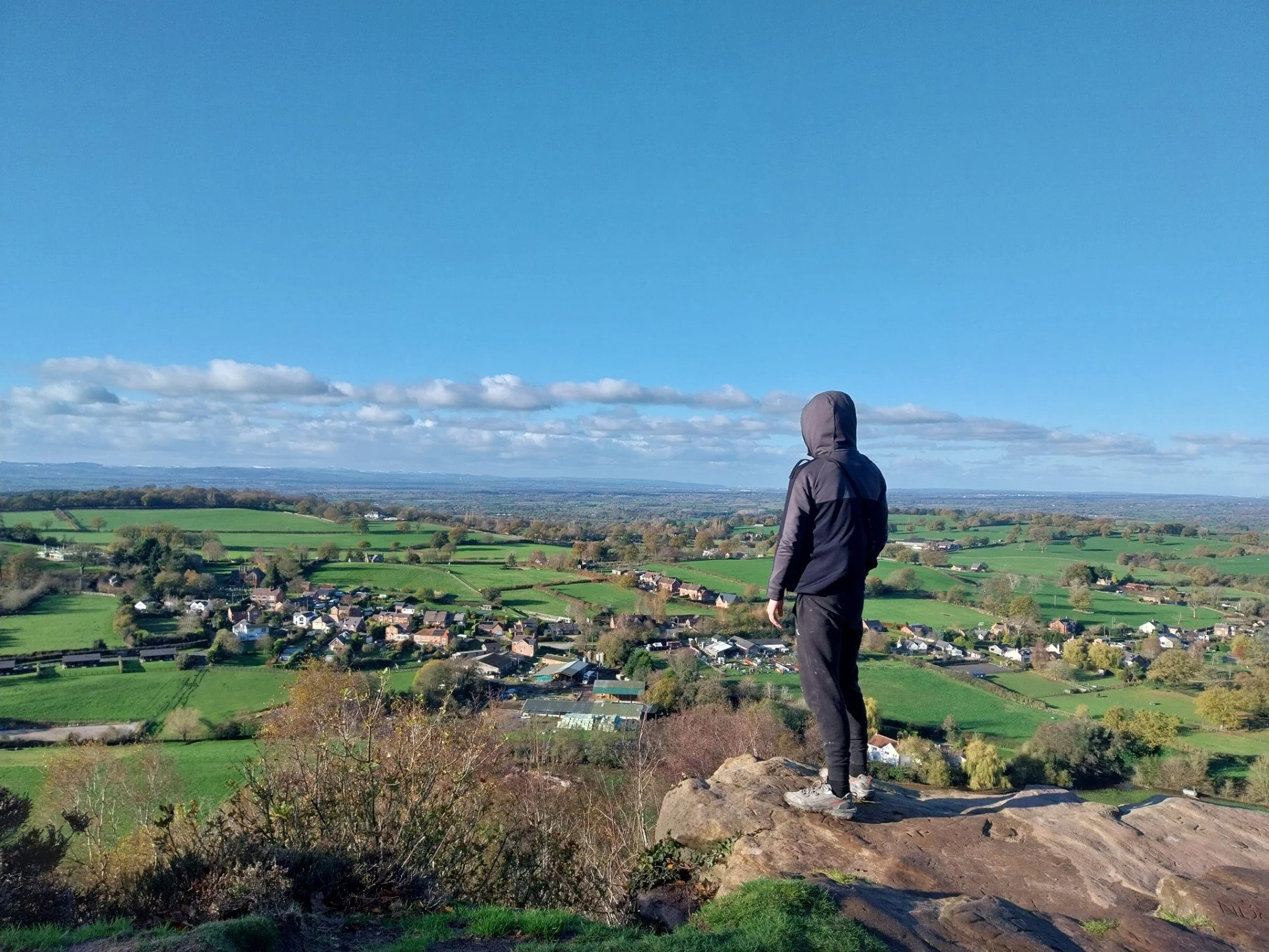 Person in a gray jacket and black pants standing on a rocky hilltop overlooking a green landscape with houses, fields, and distant hills under a blue sky with scattered clouds.