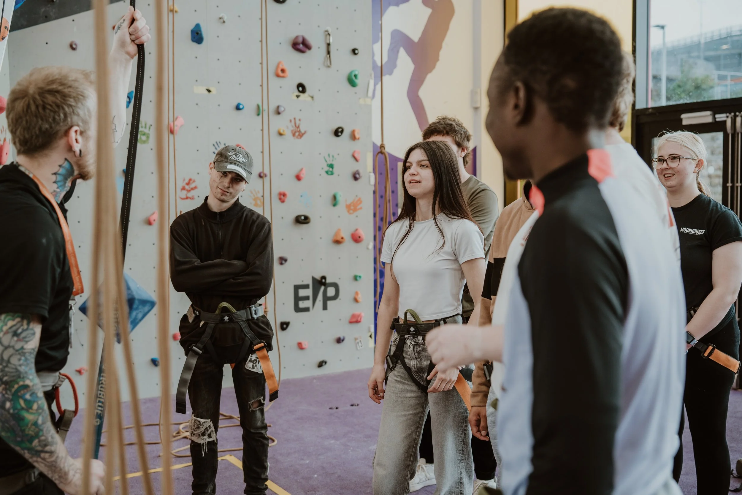 People gathered indoor at a rock climbing gym, listening to an instructor demonstrating climbing equipment.