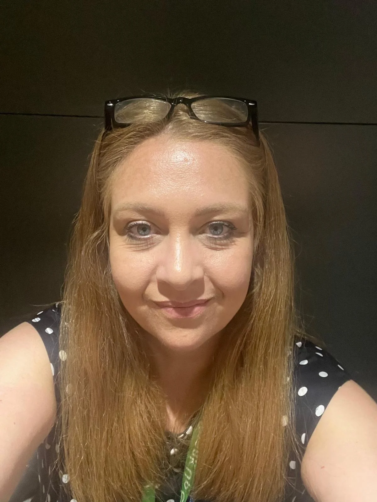 A woman with long red hair wearing glasses on her head, taking a selfie against a dark background. She is dressed in a black top with white polka dots and has a slight smile.