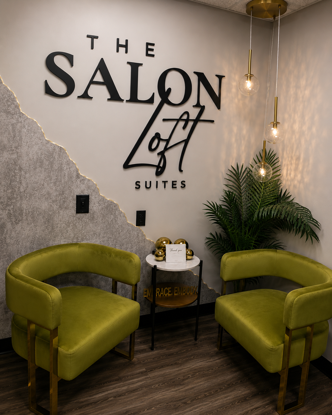 Interior of a waiting area at the Salon of Light Suites, featuring two lime green chairs, a small round table with gold decorative balls and a thank you card, a large green plant, modern hanging light fixtures, and a wall sign with the salon's name.