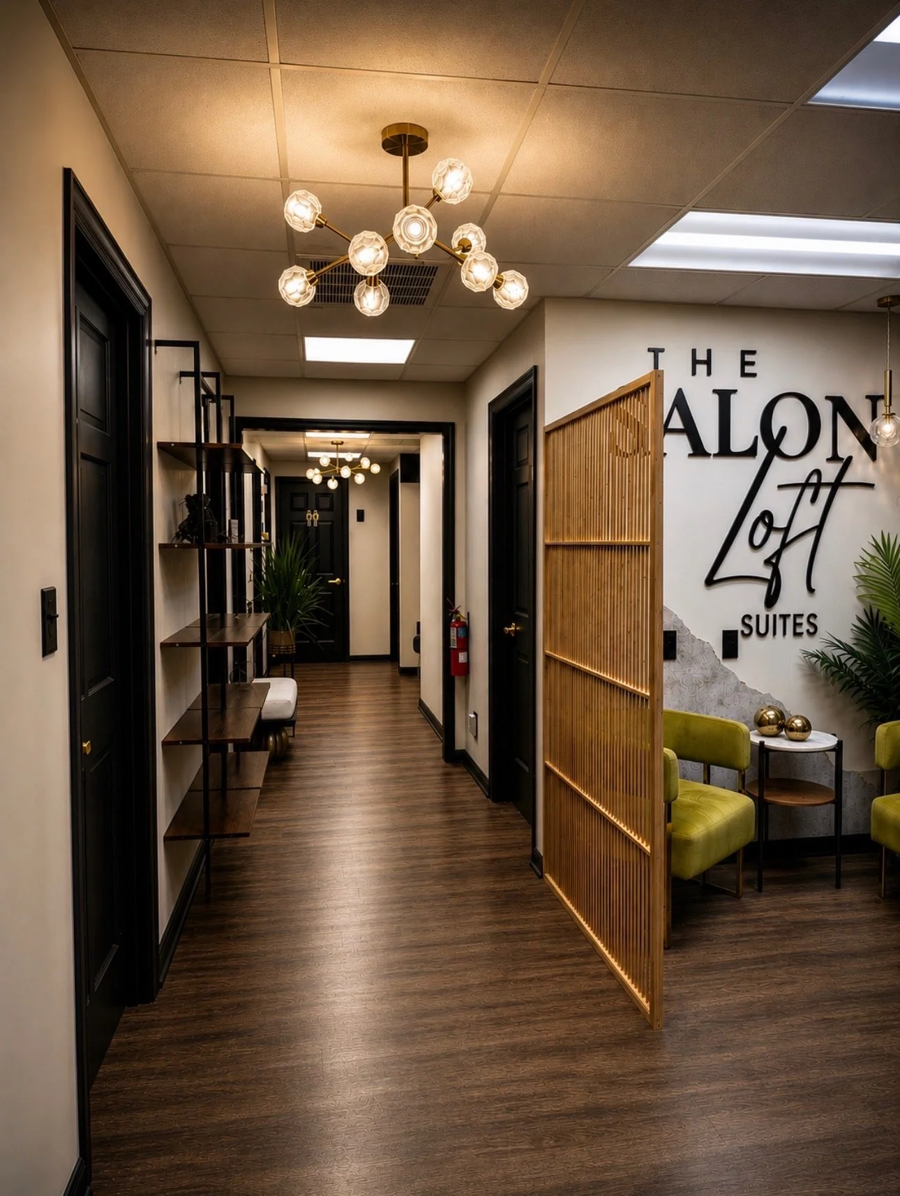Interior view of a modern, stylish office or lounge area with dark wood flooring, black doors, decorative shelves, green chairs, a small round table with gold decor, and a wall sign reading 'The Salon Loft Suites'. There are contemporary ceiling lights and potted plants.