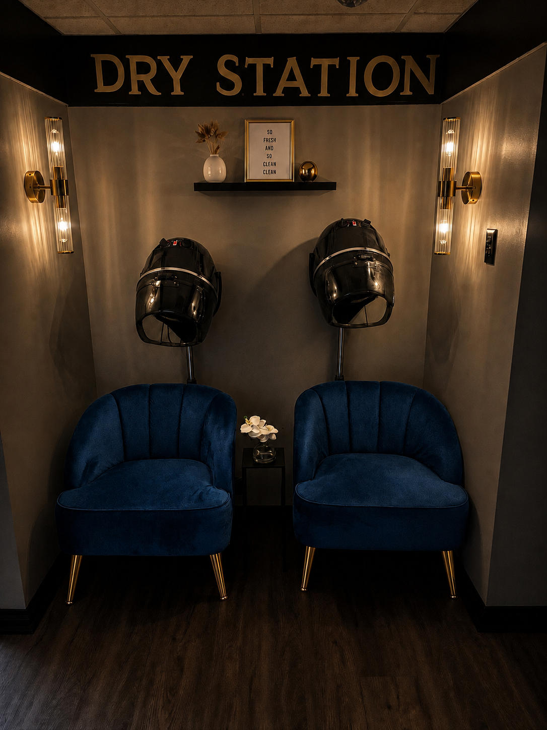 A modern salon dry station with two blue velvet chairs, hair drying stations, a small black table with a white flower in a black vase, wall sconces, and a decorative shelf with a white vase and framed sign that reads 'SO FRESH AND SO CLEAN CLEAN'.
