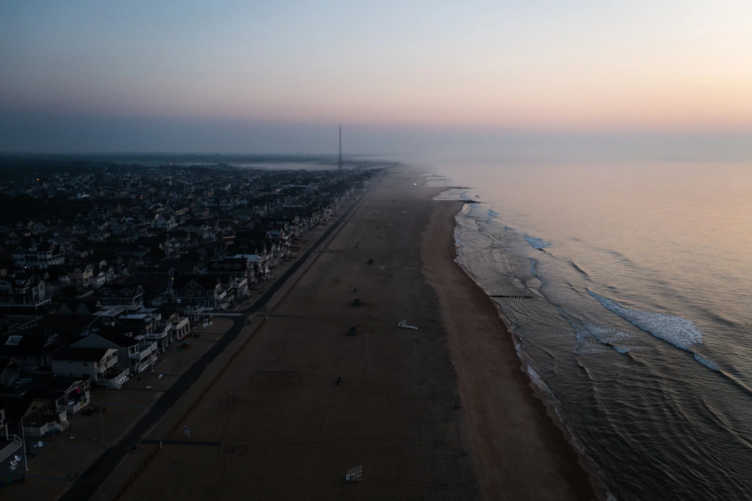 An aerial view of a coastline at dusk showing a residential area with houses along the beach, a sandy shoreline, and gentle waves in the ocean.
