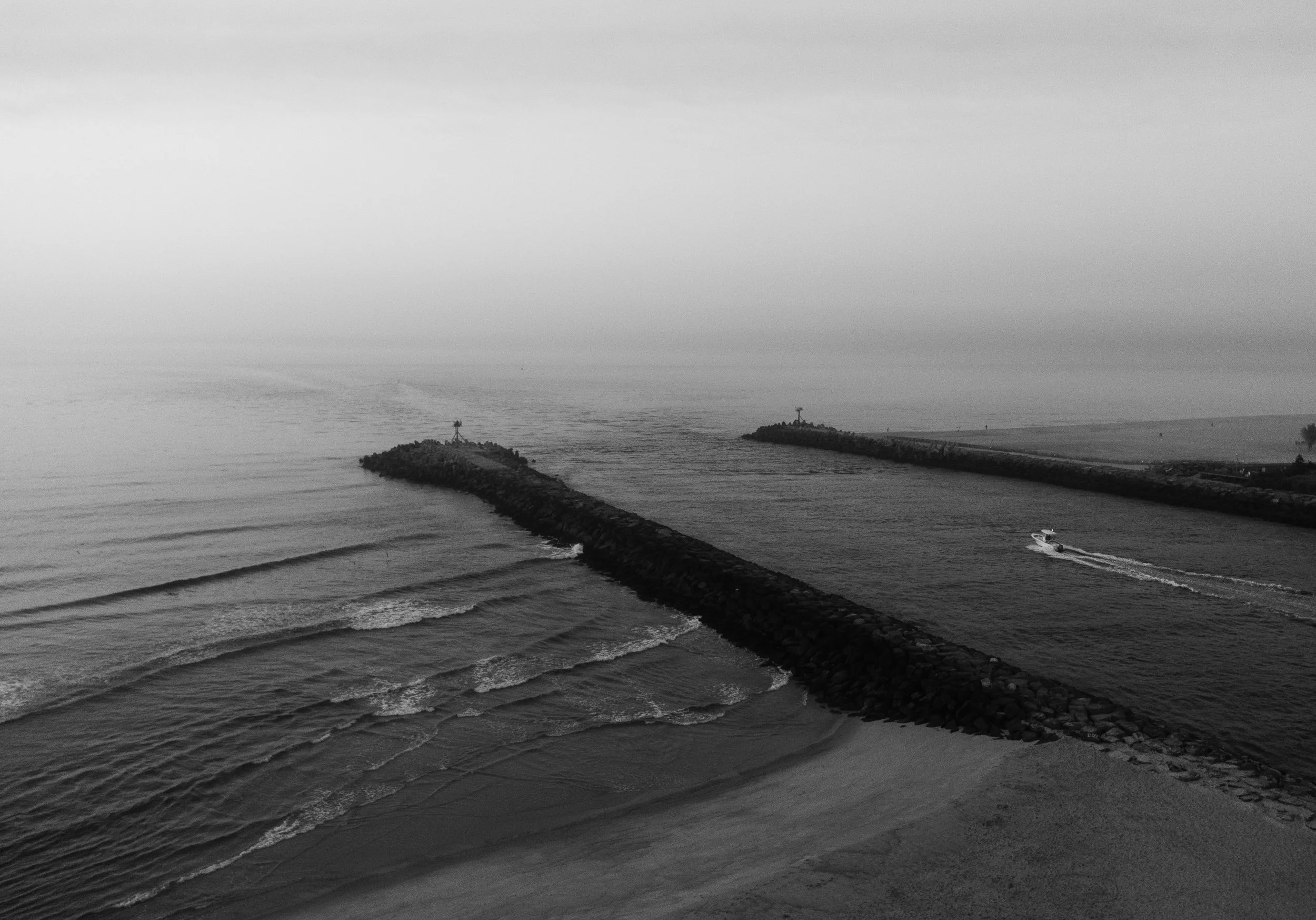 Black and white photo of a seascape with two breakwaters extending into the water, forming a V shape. A small boat is traveling near the right breakwater. The sky is overcast.