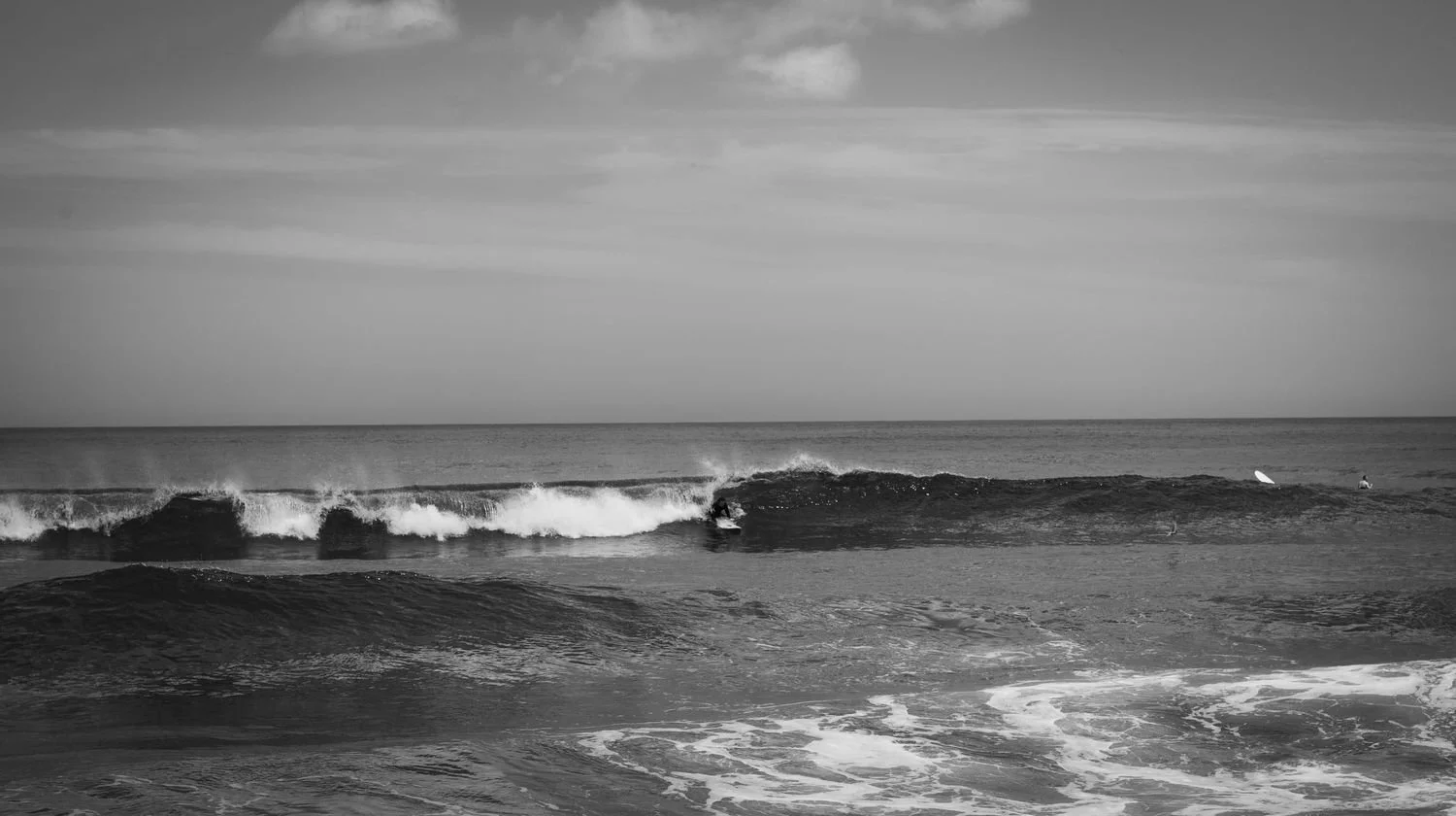 Black and white photo of a beach scene with waves, a surfer riding a wave, and a surfboard in the water.