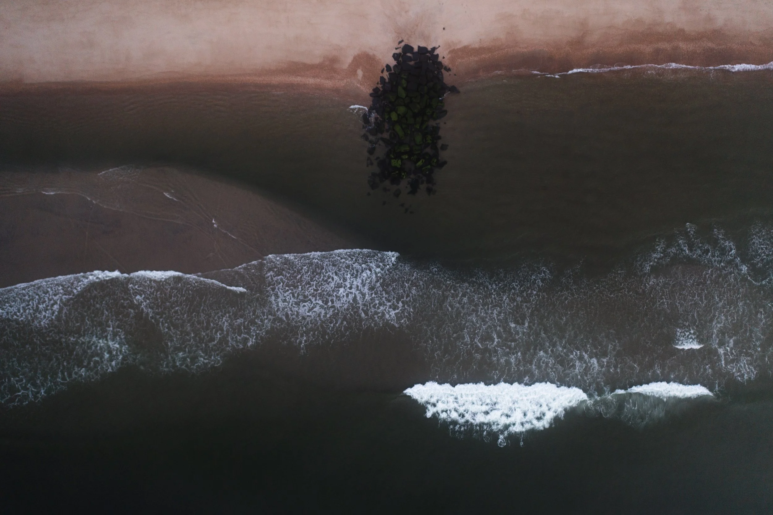 A bird's eye view of the shoreline with waves crashing onto the beach and a pile of black stones.