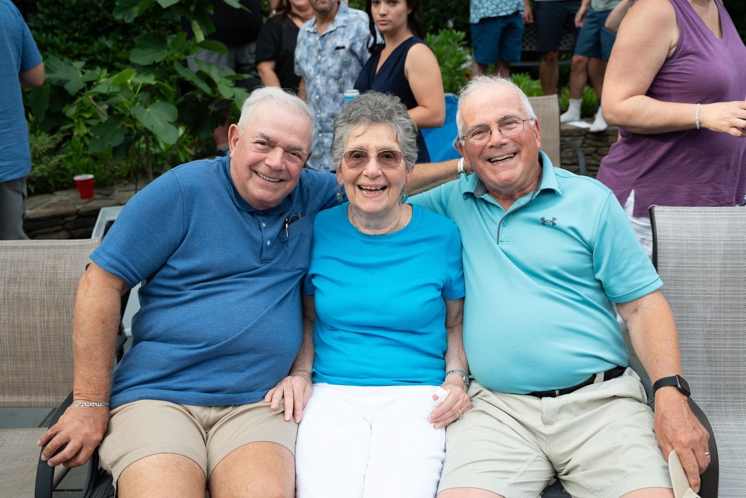 Three smiling older adults sitting closely together outdoors at a social gathering, with other people in the background.