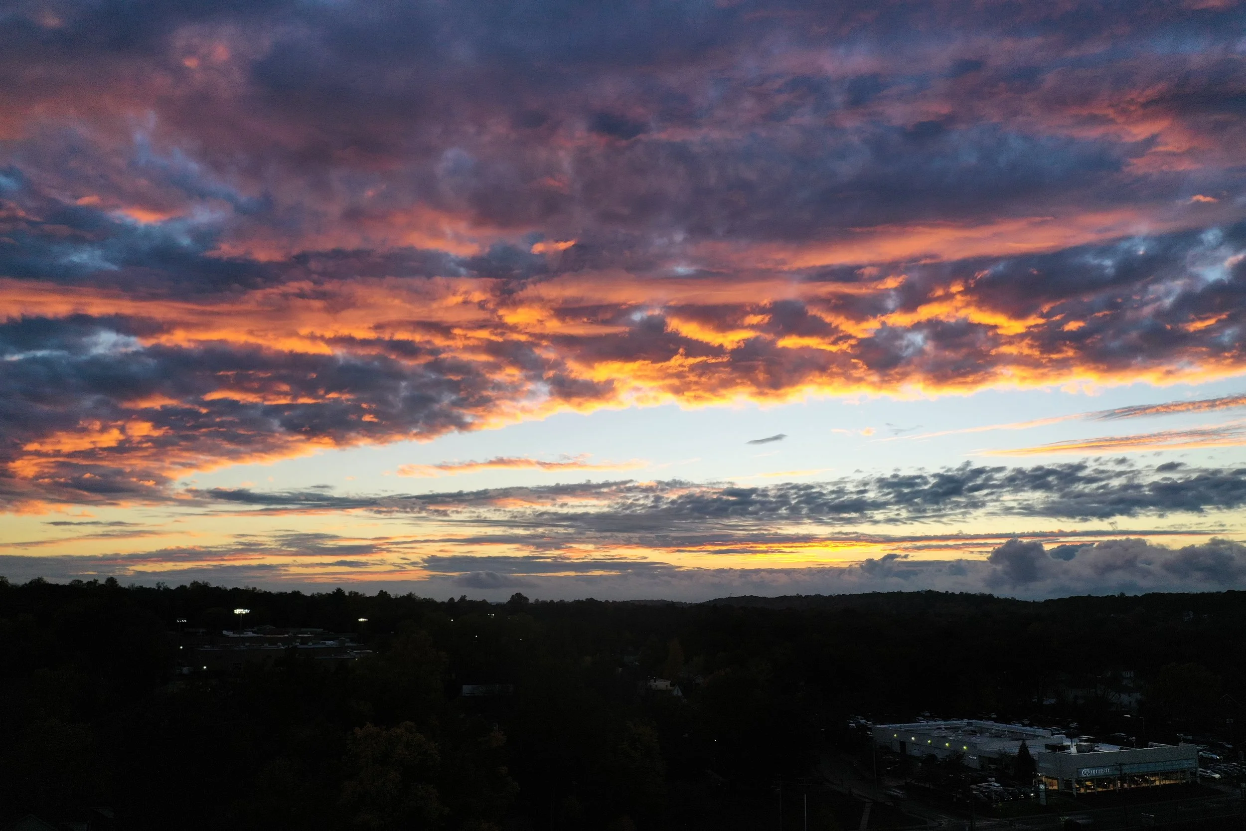 Sunset sky with dark clouds illuminated in shades of purple, pink, orange, and yellow over a landscape with trees and a few buildings.