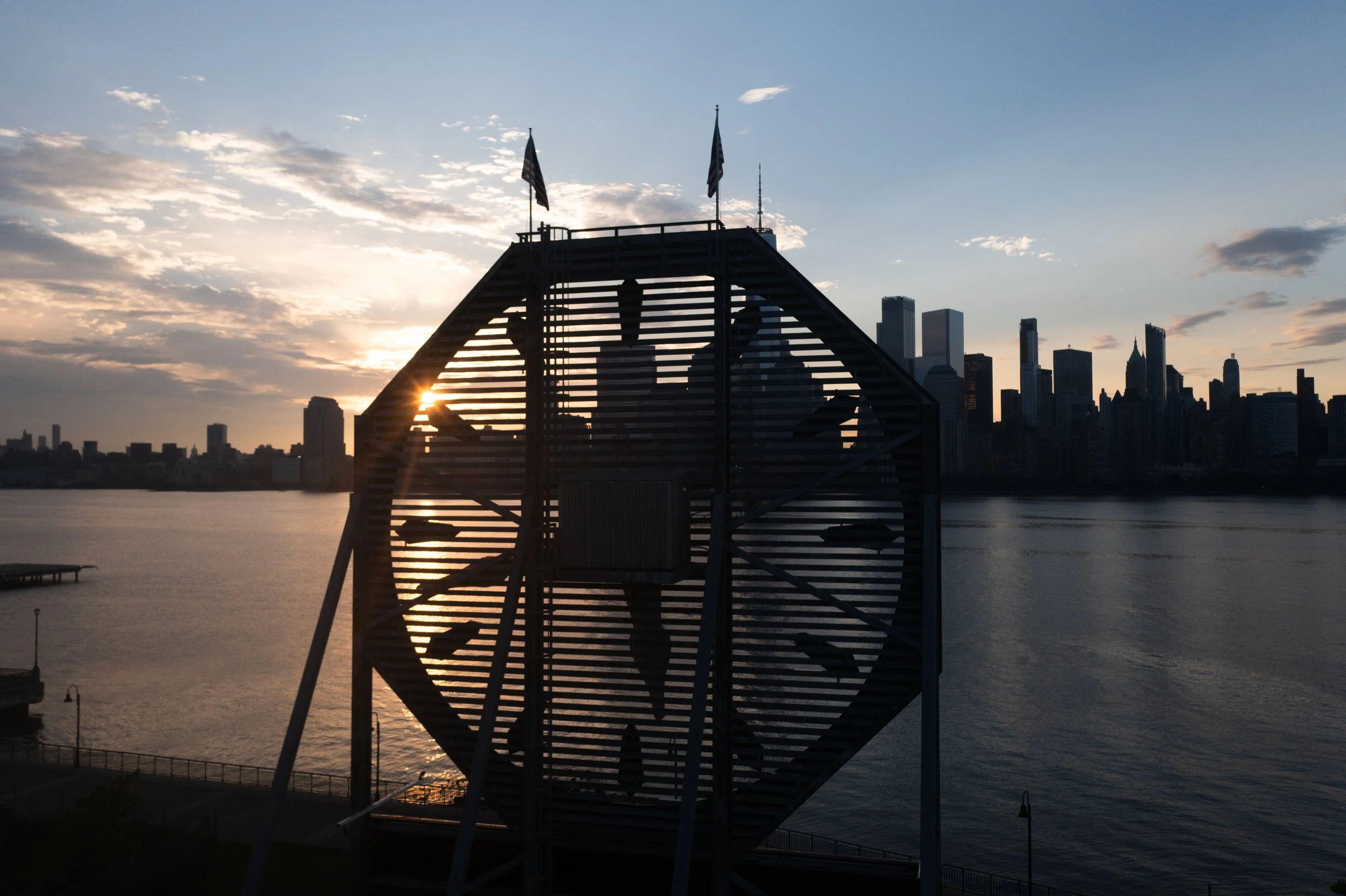Silhouette of a staircase structure with the New York City skyline in the background during sunset, with the sun partially visible through the stairs.