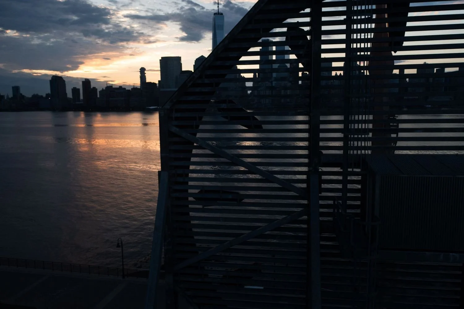City skyline at sunset with reflected water, seen through a dark metal or wooden structure in the foreground.
