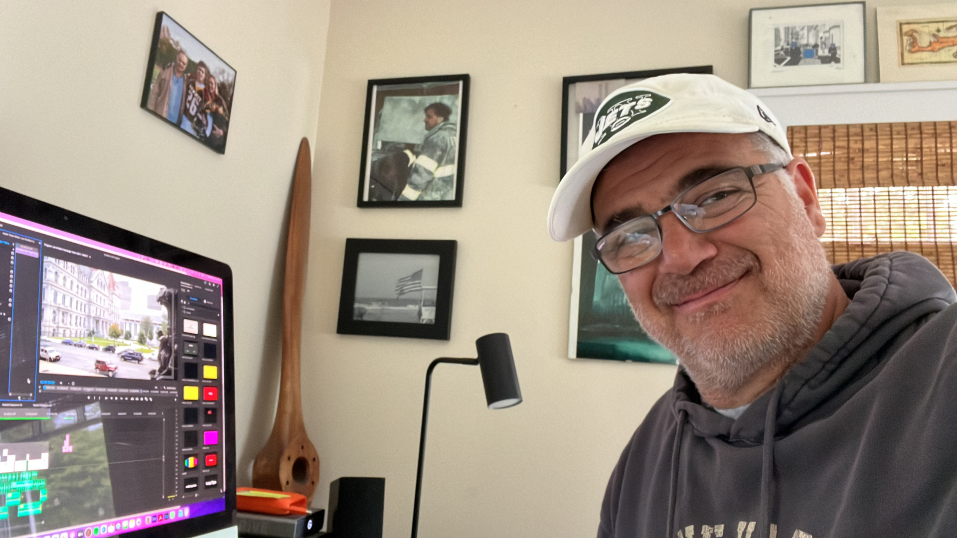 A smiling man wearing glasses, a white cap with a sports logo, and a gray hoodie, sitting in front of a computer monitor displaying video editing software. Behind him are framed pictures on the wall, a wooden paddle, and a window with bamboo blinds.