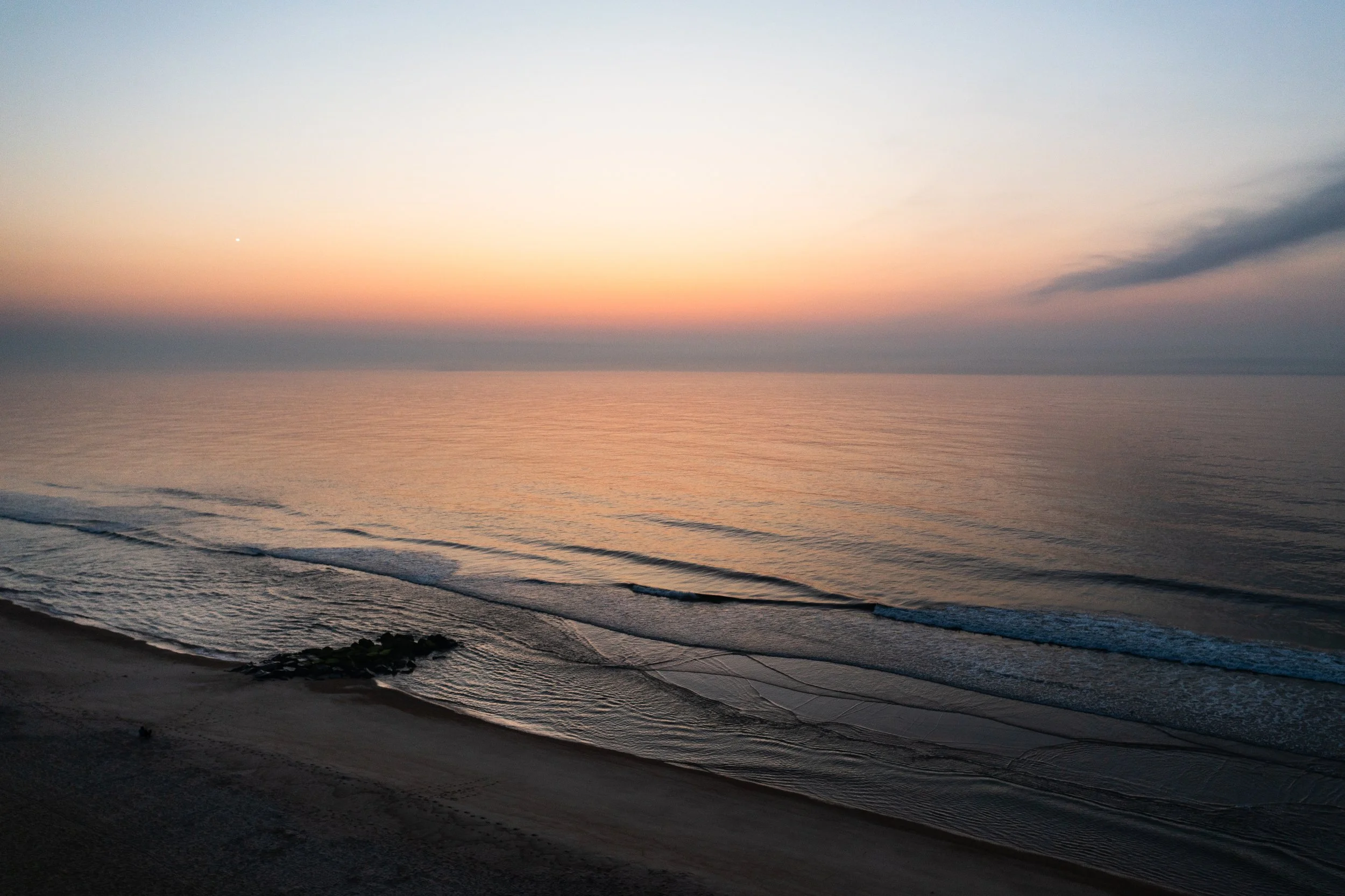 Sunset over the ocean with a pink and orange sky, gentle waves on the shore, and rocky coastline in the foreground.