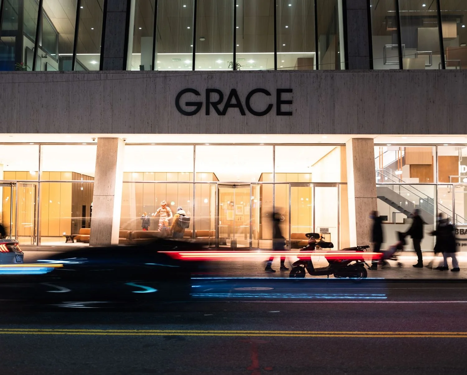 Nighttime view of a modern building with large glass windows and the name 'GRACE' on the facade, busy street with blurred motion of vehicles and pedestrians in front, and downtown city lights.