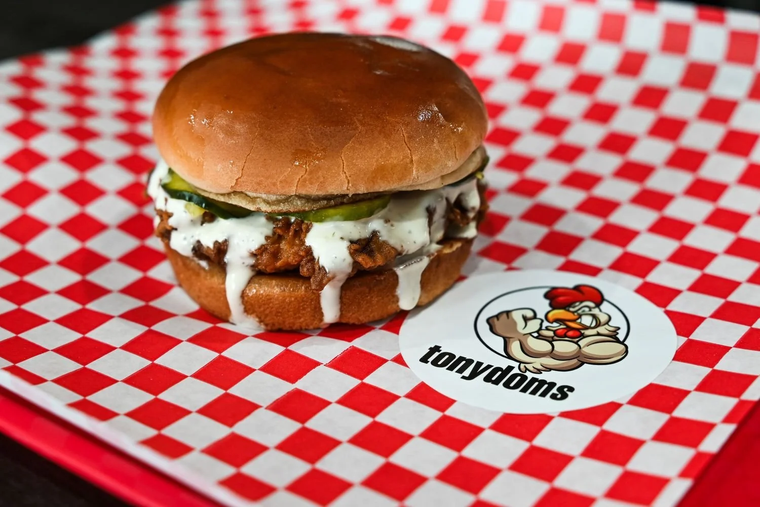 Close-up of a burger with pickles, shredded meat, and white sauce on a checkered paper with a cartoon chicken logo and the word 'tonydöms'.