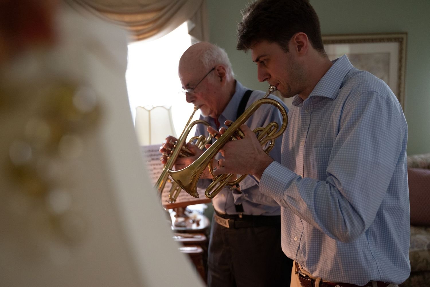 Two men playing trumpets indoors, focused on their music, with sheet music on a stand in front of them.