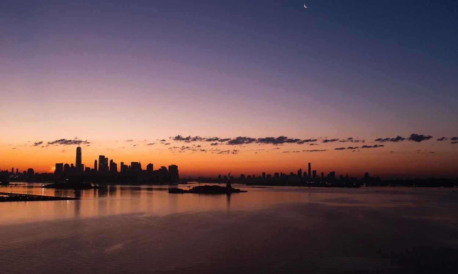 City skyline at sunset with the Statue of Liberty in the foreground, reflecting on the water, with a clear sky and a crescent moon.