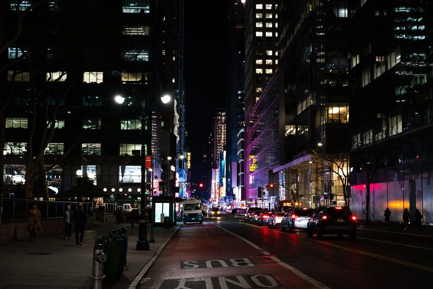 Nighttime city street scene with illuminated buildings, neon lights, cars, and people walking on the sidewalk.