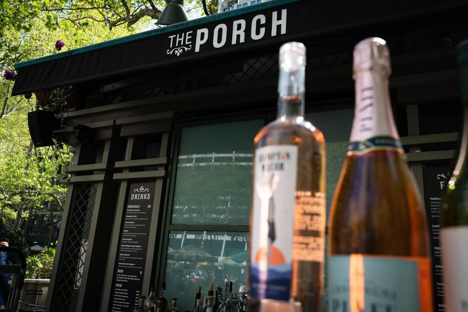Outdoor scene of a bar called 'The Porch' with bottles of alcohol in foreground, a menu on the wall, and trees with green leaves in the background.
