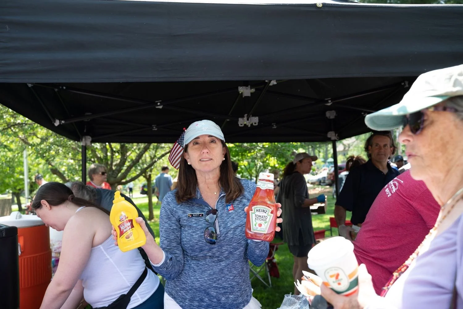 A woman in a blue long-sleeve shirt holding bottles of ketchup and mustard at an outdoor gathering, under a black canopy, with people in the background and trees around.