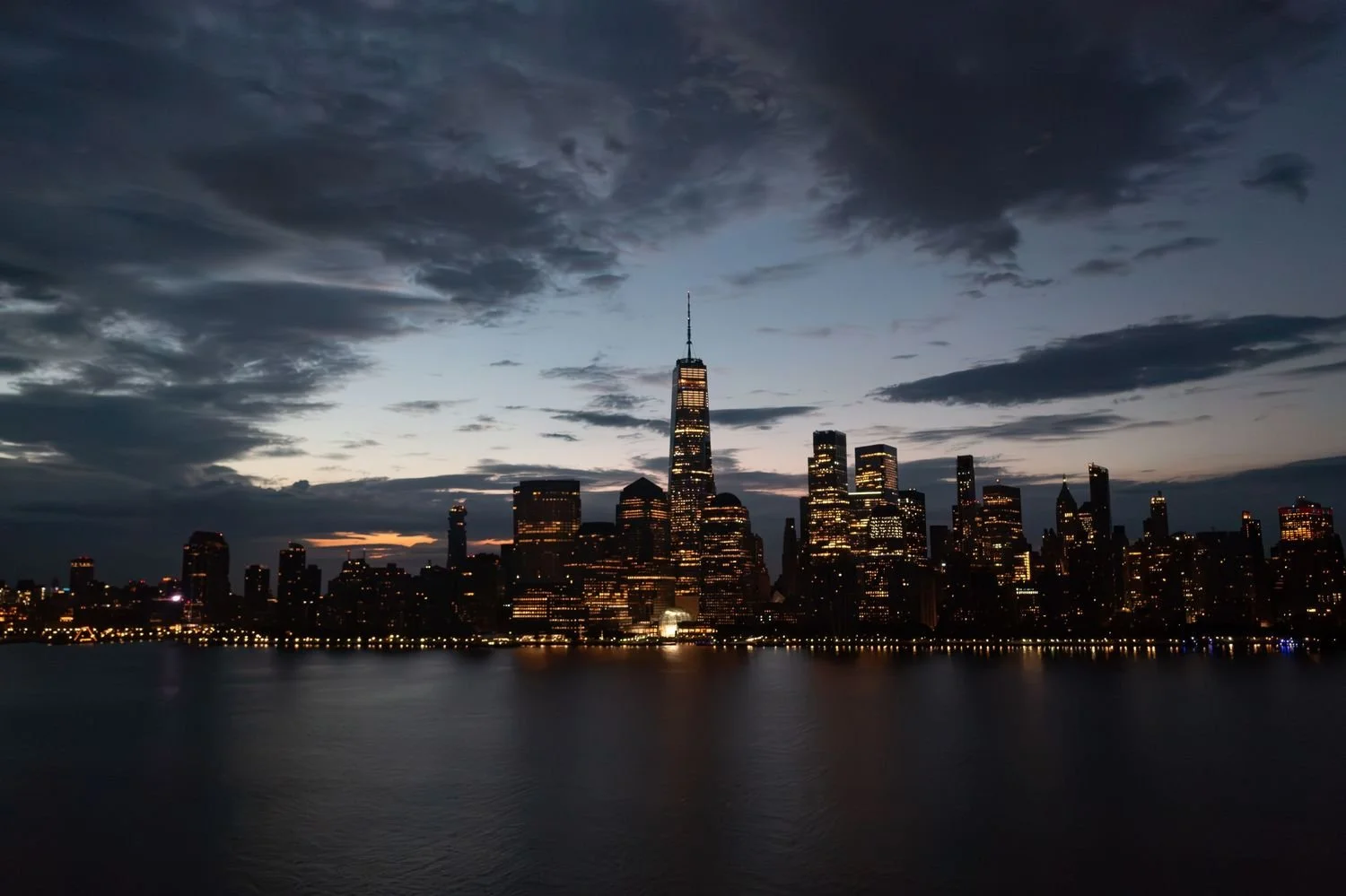 Nighttime city skyline of Manhattan, New York City, with One World Trade Center illuminated, reflecting on the water.
