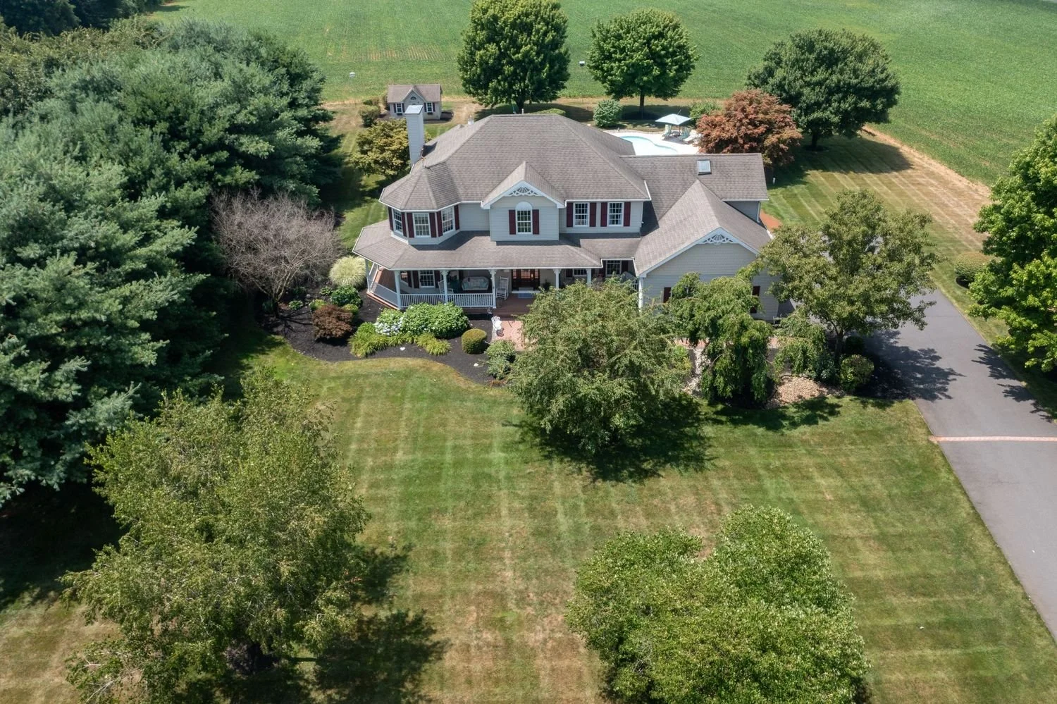 A large, two-story suburban house with a front porch, surrounded by green trees, a well-maintained lawn, and a driveway on the right side, in a spacious yard with a garden and a swimming pool.