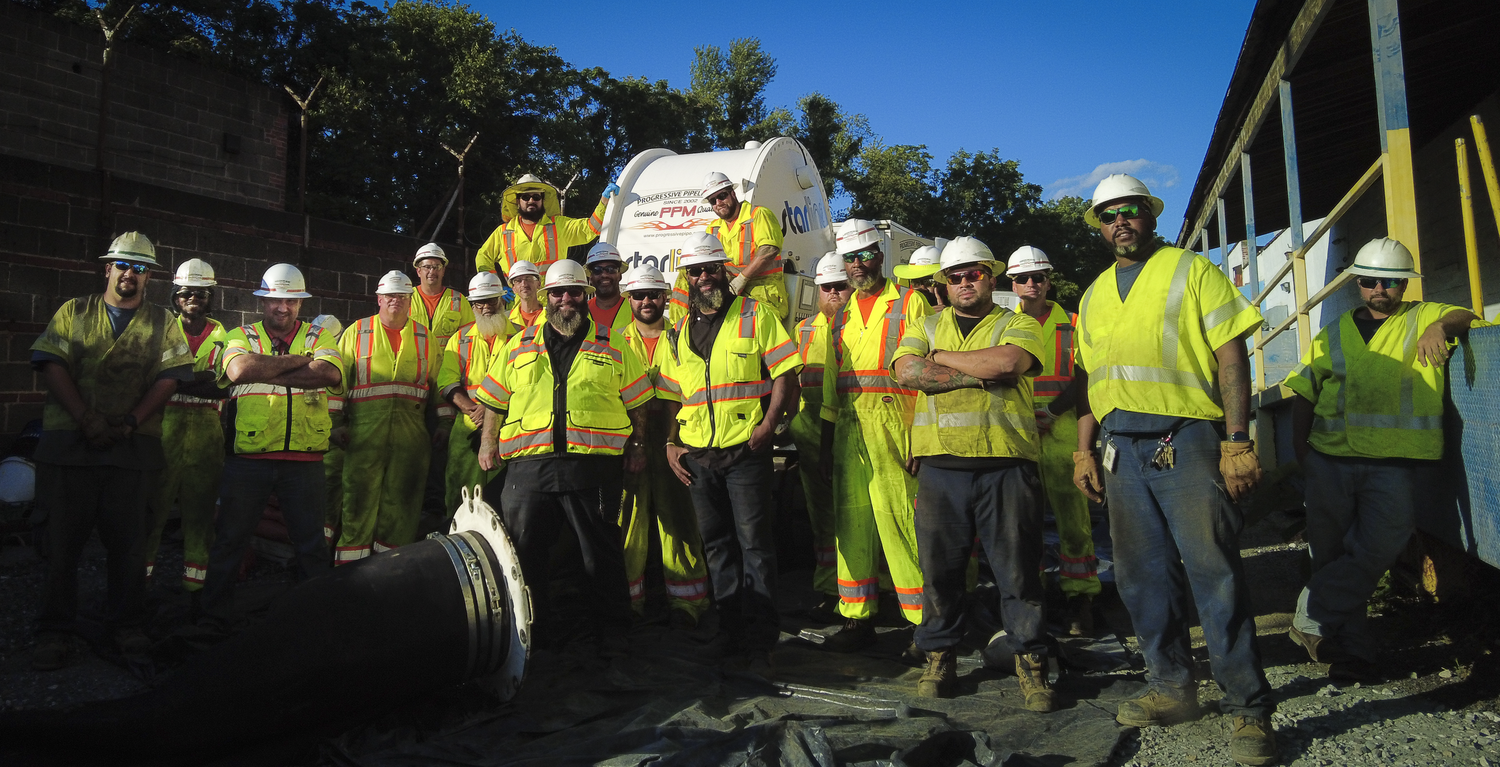 Group of construction workers in safety vests and hard hats at a construction site. Some are standing near a large pipe component, with equipment and a building under construction visible in the background.
