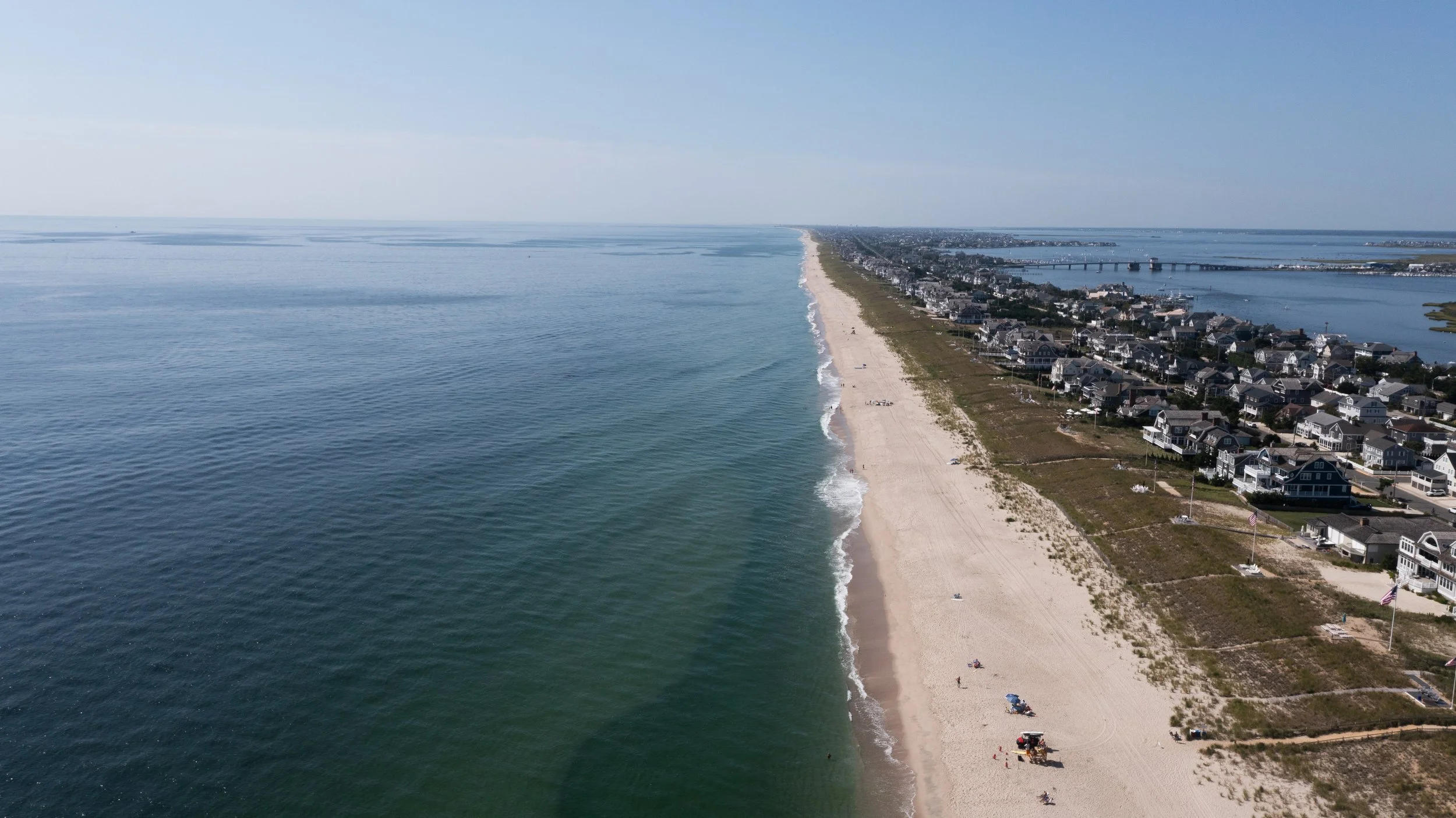 An aerial view of a long sandy beach with calm ocean waves on the left and a row of houses on the right, separated by a strip of land, under a clear blue sky.