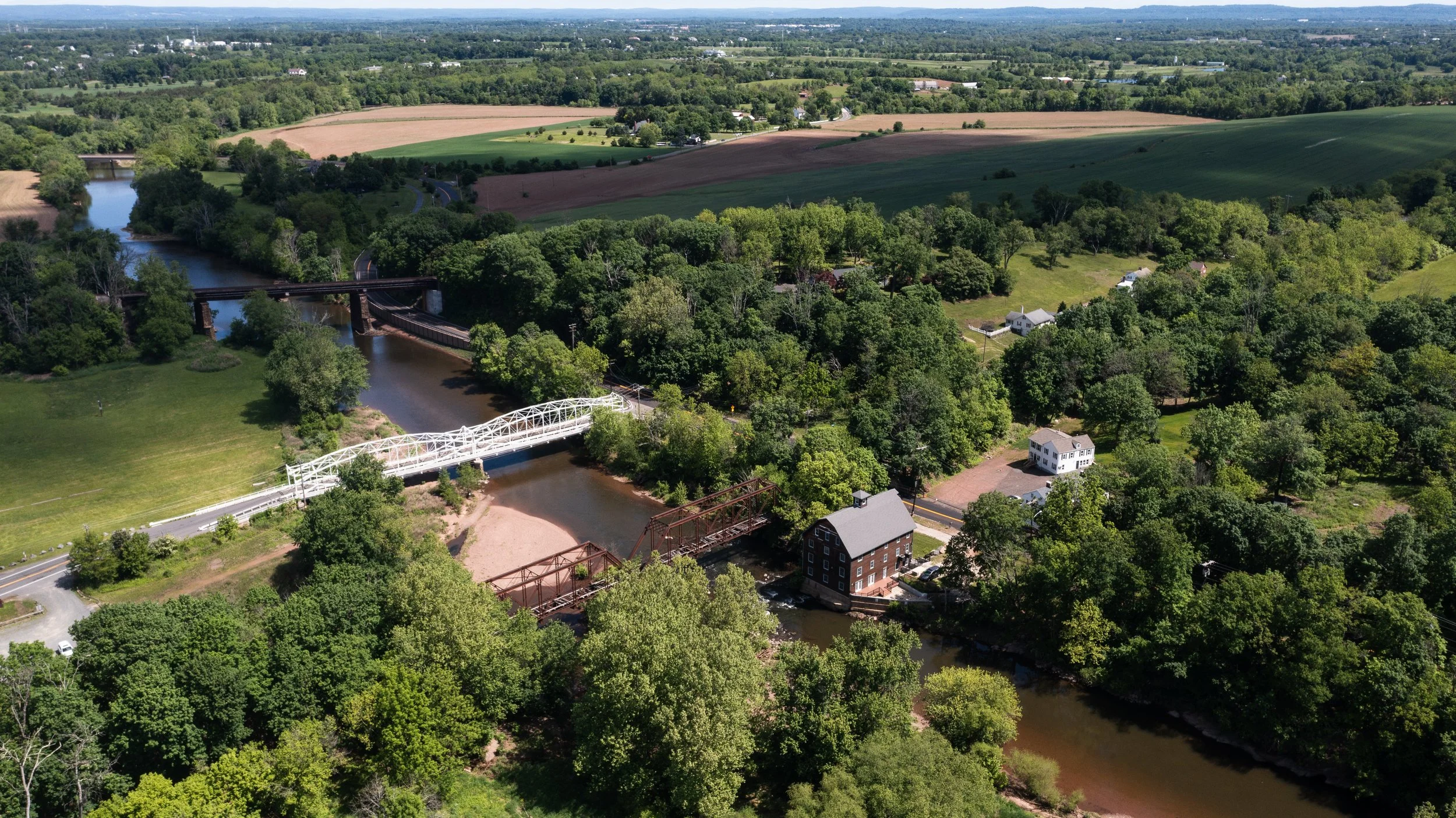 Aerial view of a rural landscape with trees, a river, bridges, houses, and farmland under a clear sky.
