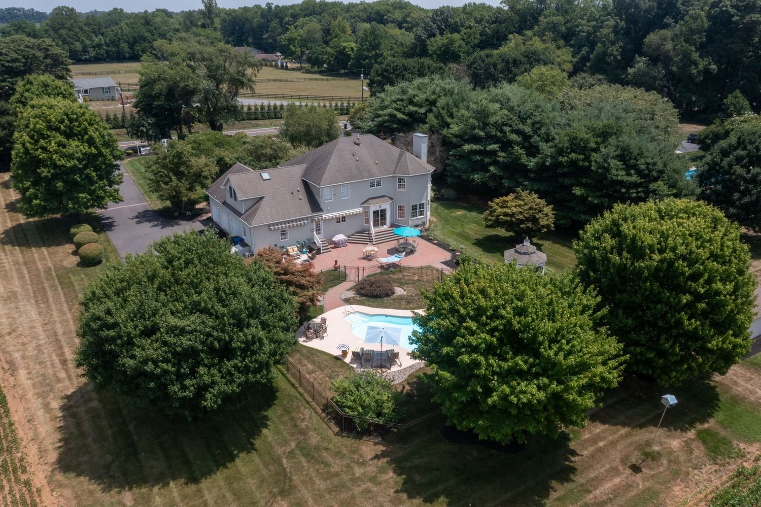 A large gray house with a backyard featuring a swimming pool, patio furniture, trees, and a well-manicured lawn, set in a suburban area surrounded by other houses and greenery.