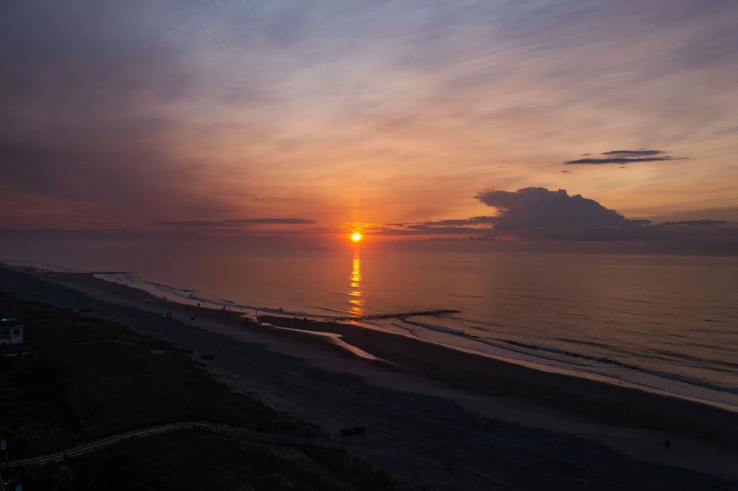 Sunset over the ocean viewed from a beach with a few people, gentle waves, and a partly cloudy sky with purple, orange, and blue hues.